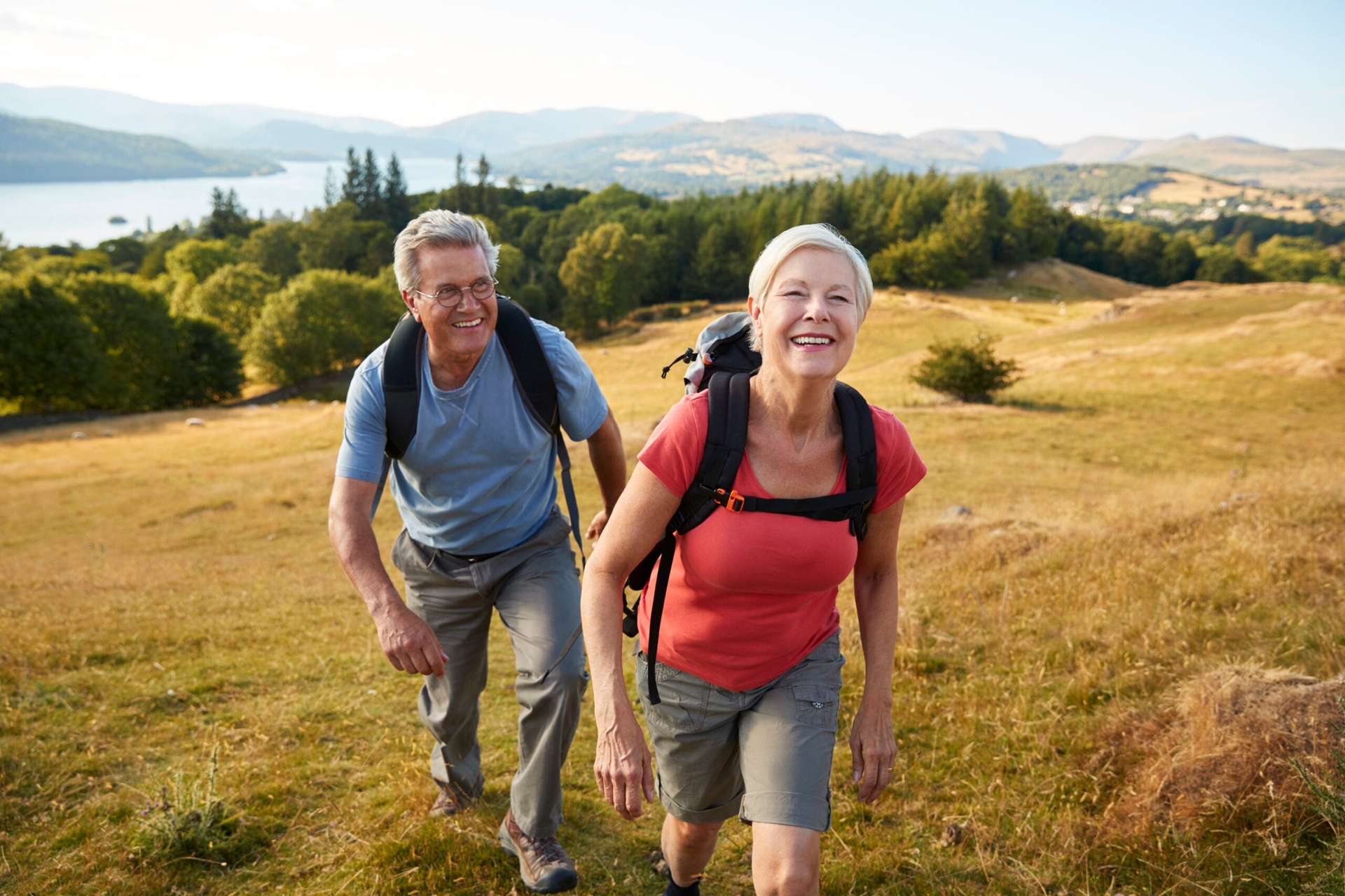 Two elderly people with backpacks smiling as they hike up a grassy hill with a scenic view of trees, a lake, and mountains in the background.