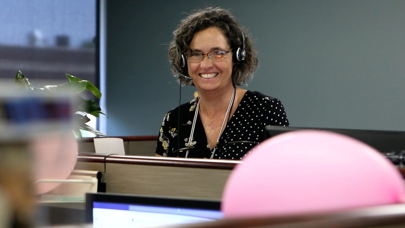 A woman with curly hair and glasses is smiling while sitting at an office cubicle, wearing a headset. A pink balloon is visible in the foreground.