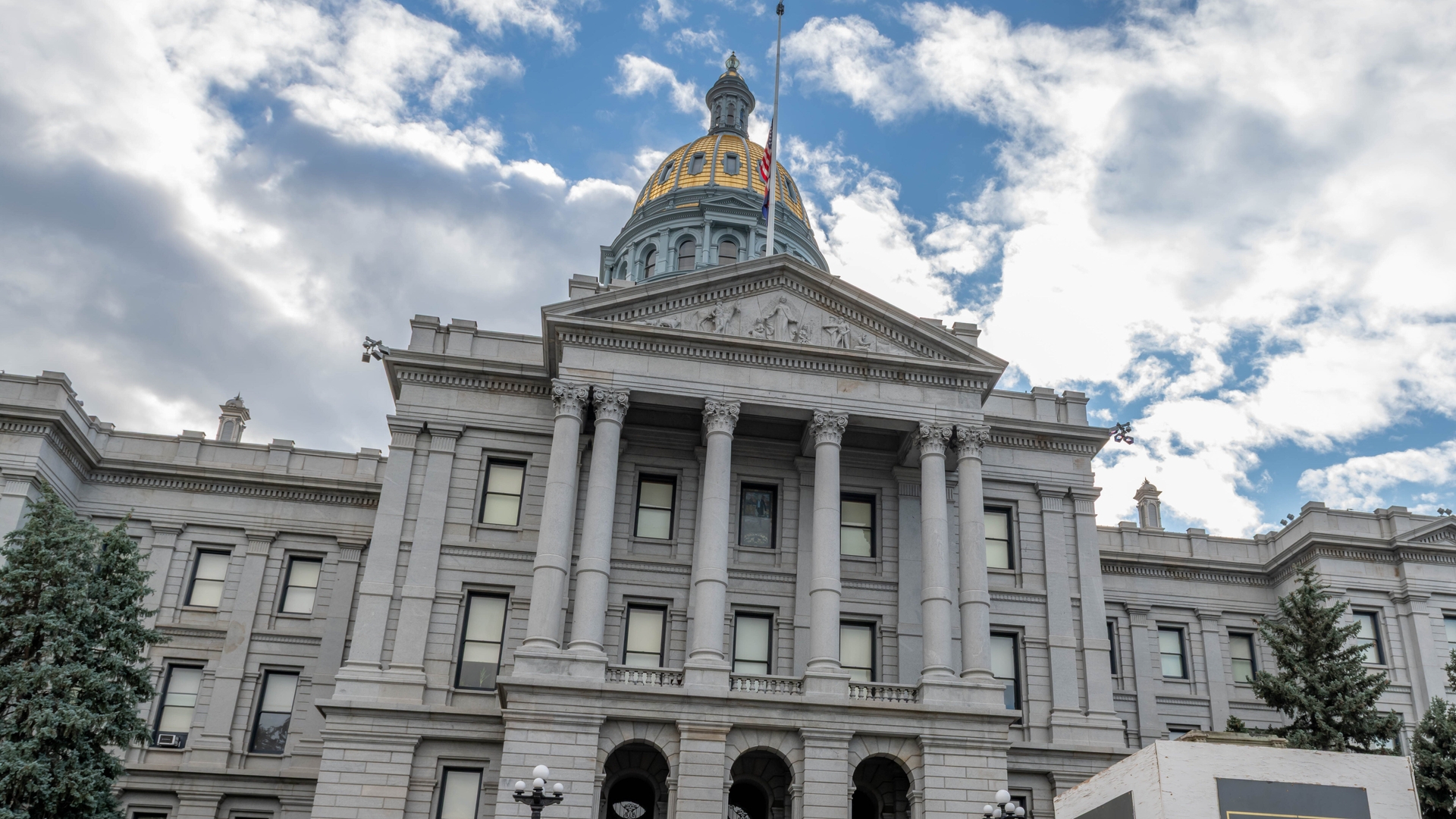 The Colorado State Capitol building in Denver with a gold-domed roof, columns, steps, and a stone marker in front under a partly cloudy sky.