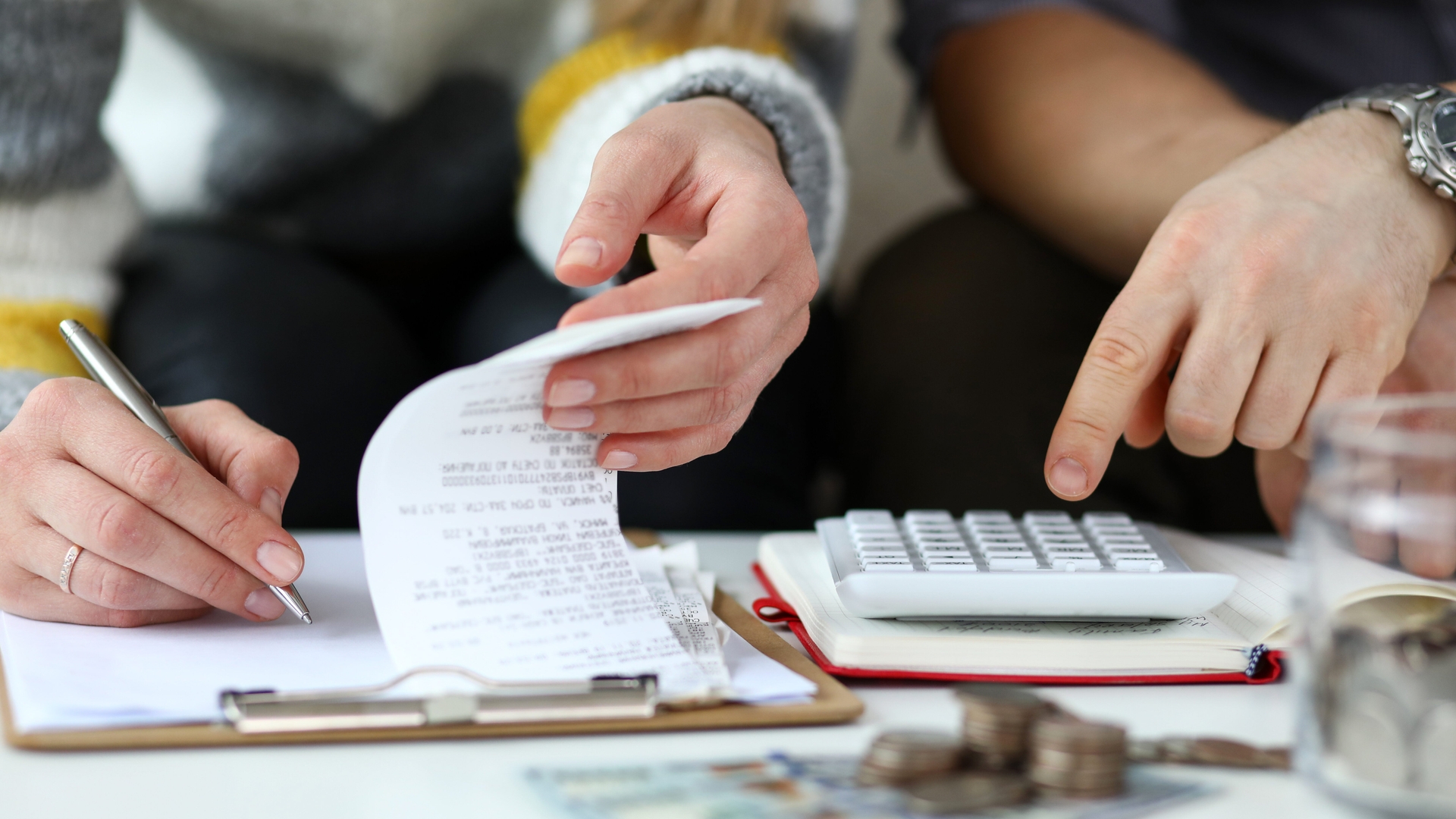 Two people review a receipt and use a calculator amid coins, bills, and documents on the table.