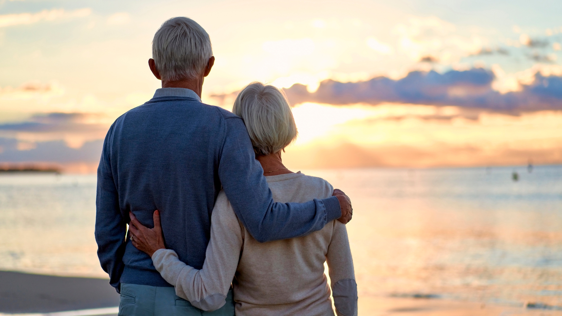 A gray-haired couple on a beach, looking away from the camera and toward the setting sun.