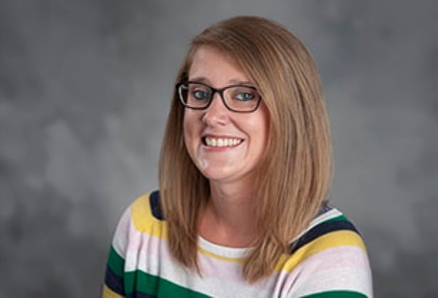 Woman with straight light brown hair and glasses, wearing a striped sweater, smiling in front of a gray background.