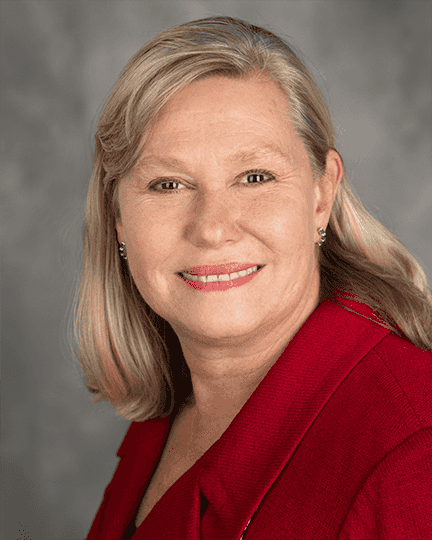 A middle-aged woman with shoulder-length blonde hair wearing a red blazer smiles at the camera against a gray background.