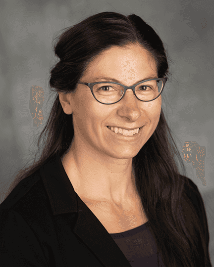 Portrait of a woman with long dark hair, wearing glasses, a dark blazer, and a slight smile, against a neutral background.