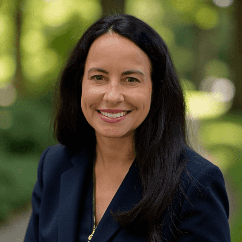 A woman with long dark hair and a navy blazer smiles outdoors with trees and greenery in the background.