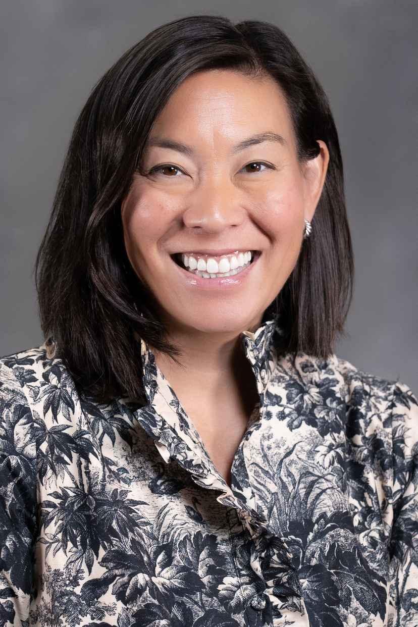 A woman with straight, shoulder-length dark hair is smiling at the camera. She is wearing a patterned blouse with a floral design and stud earrings, set against a plain gray background.