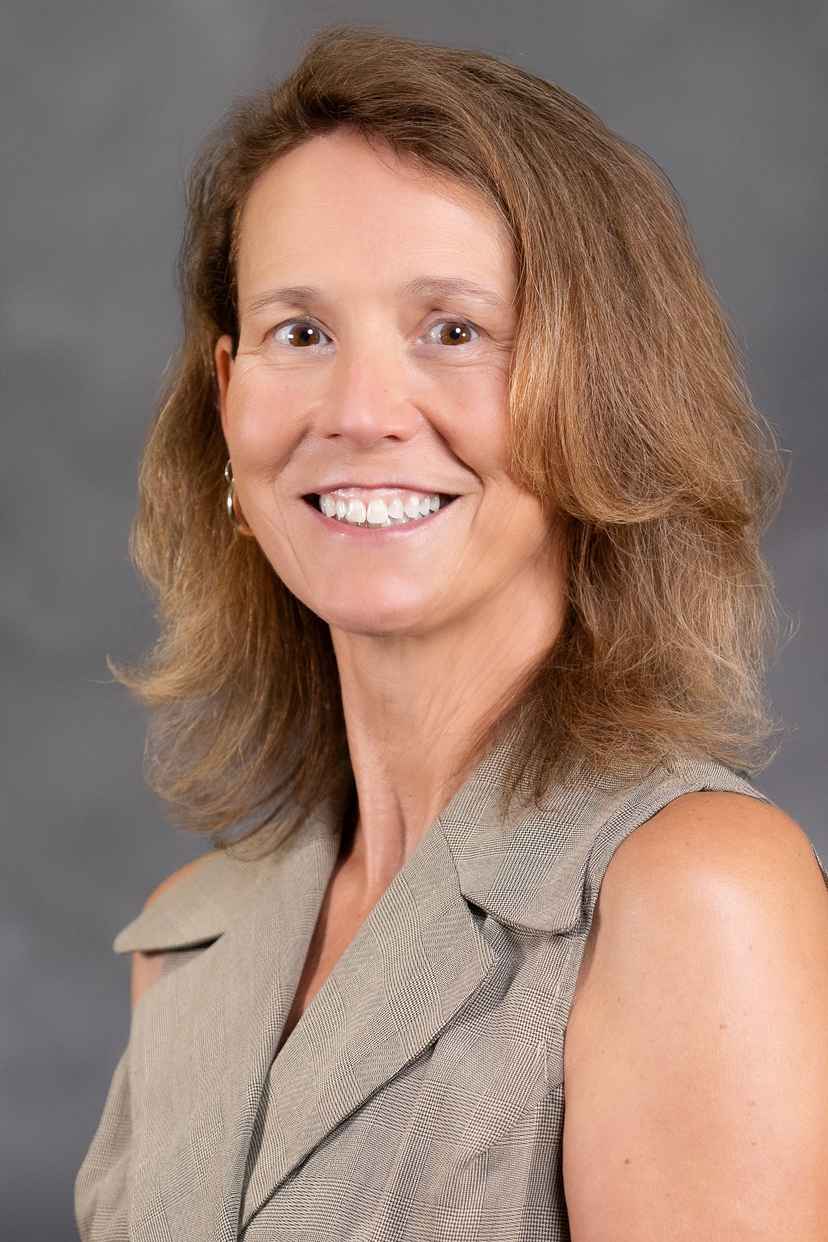 A woman with shoulder-length light brown hair, wearing a sleeveless beige top, smiles in front of a gray background.