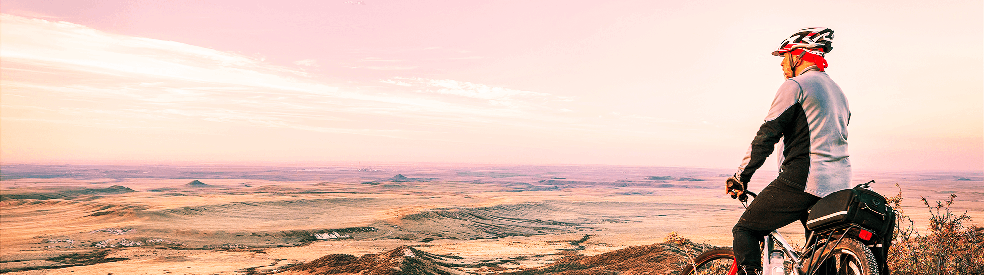 A person wearing a helmet and cycling gear stands beside a bicycle on a hill, overlooking a vast, open landscape at sunset.
