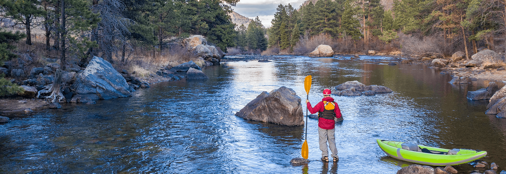 A person in outdoor gear stands with a yellow paddle beside a green kayak in a rocky, forested river scene.