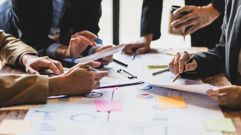 A group of people work together at a table covered with charts, graphs, tablets, and colorful sticky notes.