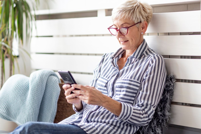 Older woman with short blonde hair and red glasses, wearing a striped shirt, sits indoors on a sofa, smiling while using her smartphone.