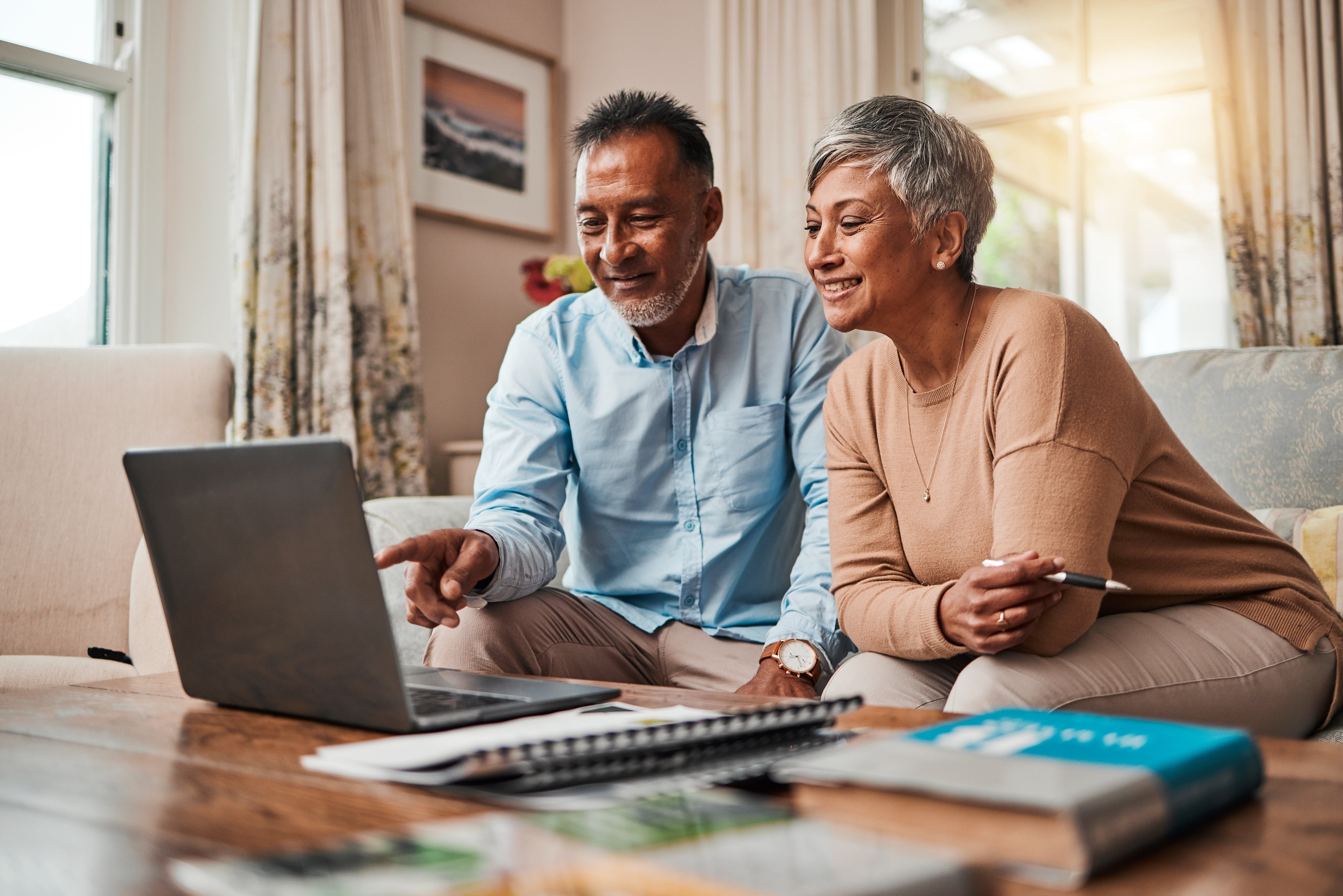 An older couple sits on a couch together, looking at a laptop screen and smiling, with paperwork and books spread out on the coffee table in front of them.