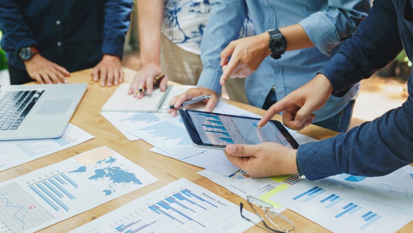 Four people review charts and graphs on paper and a tablet at a desk, with a laptop and eyeglasses nearby.