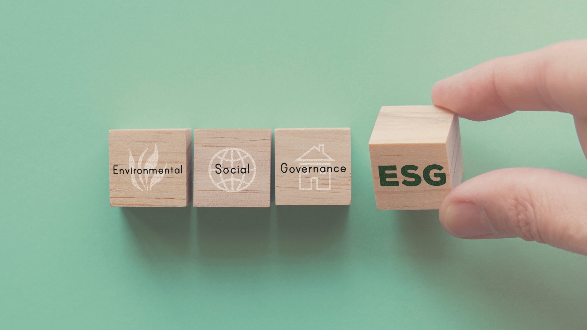 A hand holding a wooden block labeled "ESG" next to three other blocks labeled "Environmental," "Social," and "Governance" against a green background.