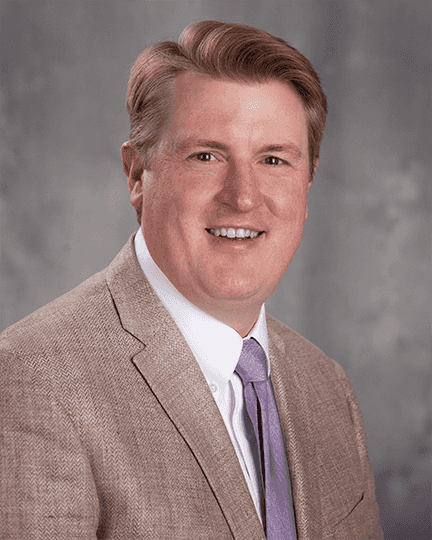 A man with light hair wearing a beige suit jacket, white shirt, and lavender tie, smiling at the camera in front of a gray background.