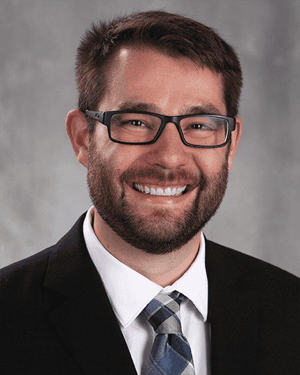 A man with glasses, a beard, and short hair smiles at the camera. He is wearing a black suit, white shirt, and a blue plaid tie, with a neutral gray background.