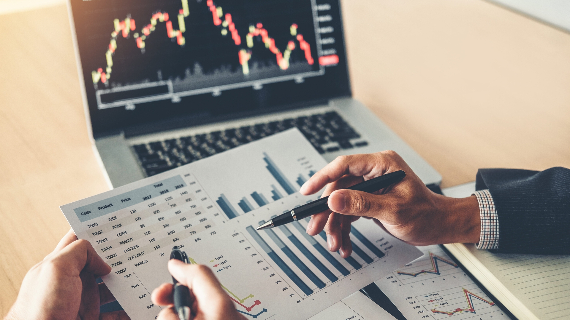 Two people analyzing financial charts and graphs on paper, with a laptop displaying a stock market graph in the background.