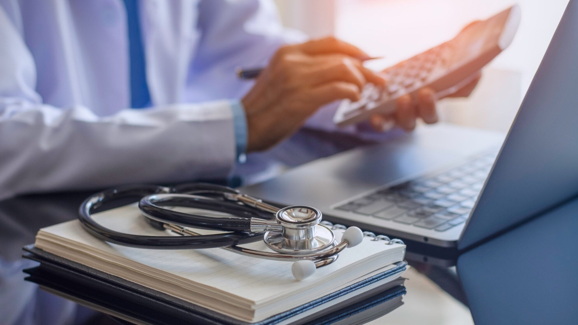 A medical professional uses a calculator and laptop with a stethoscope and notebook on the desk next to them