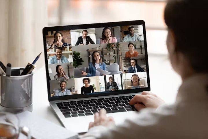 A person participates in a video conference on a laptop, displaying twelve people in a grid layout. The desk has a pen holder and a notebook.