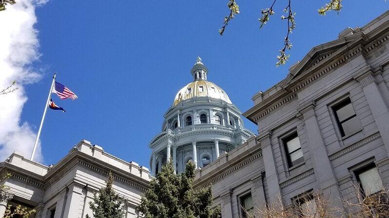The Colorado State Capitol building taken from a low angle looking up. The American and Colorado flag can be seen waving in the wind.