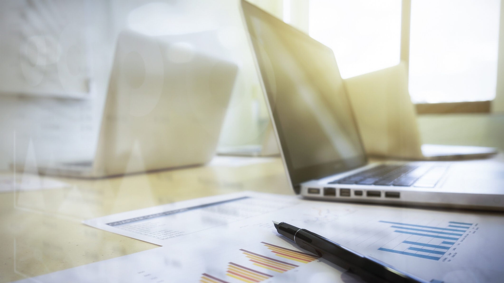 A close up of papers on a desk with two laptops sitting in the background.