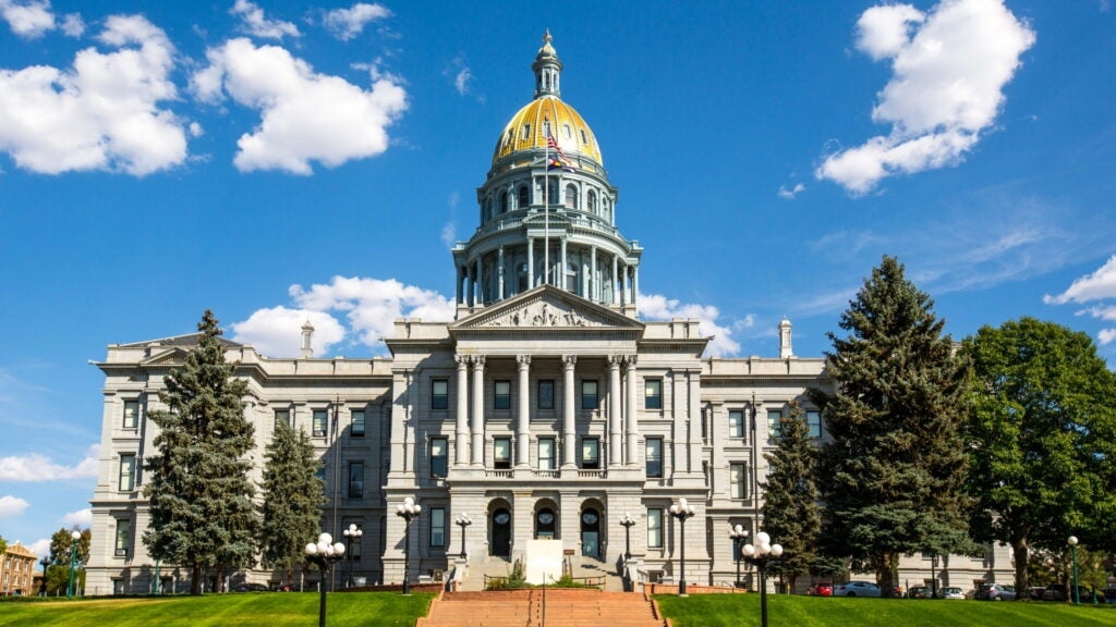 A wide shot of the Colorado State Capitol building with its golden dome, surrounded by trees and set against a bright blue sky with scattered clouds.