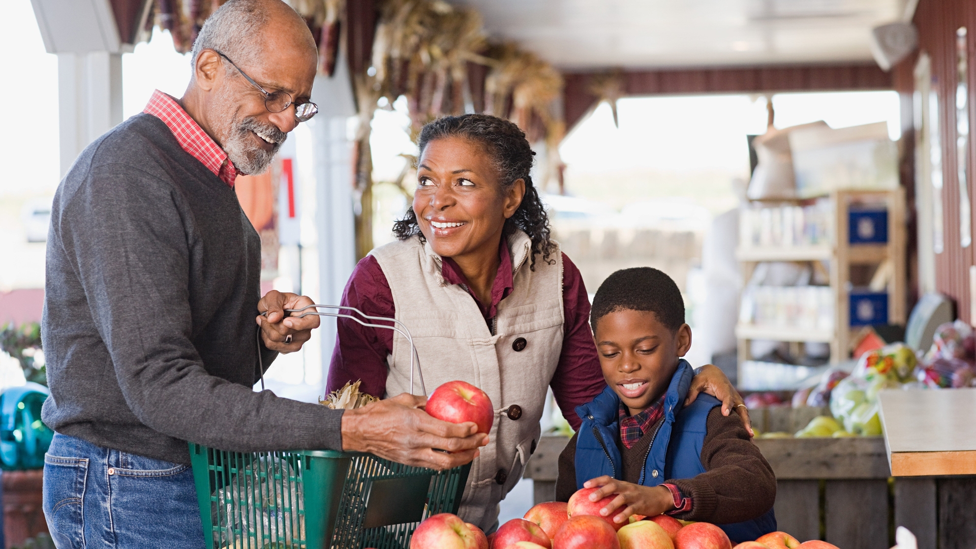 An elderly man, woman, and young boy select apples at a market stand, with the man holding a green basket and the woman and boy smiling.