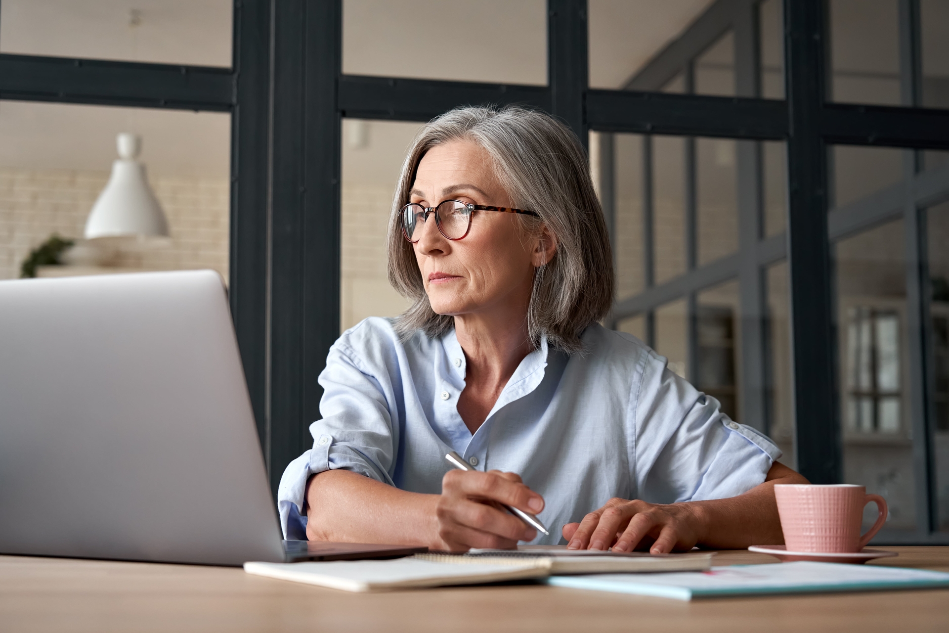 Older woman sitting at a desk looking at a laptop while holding a pen over paper.