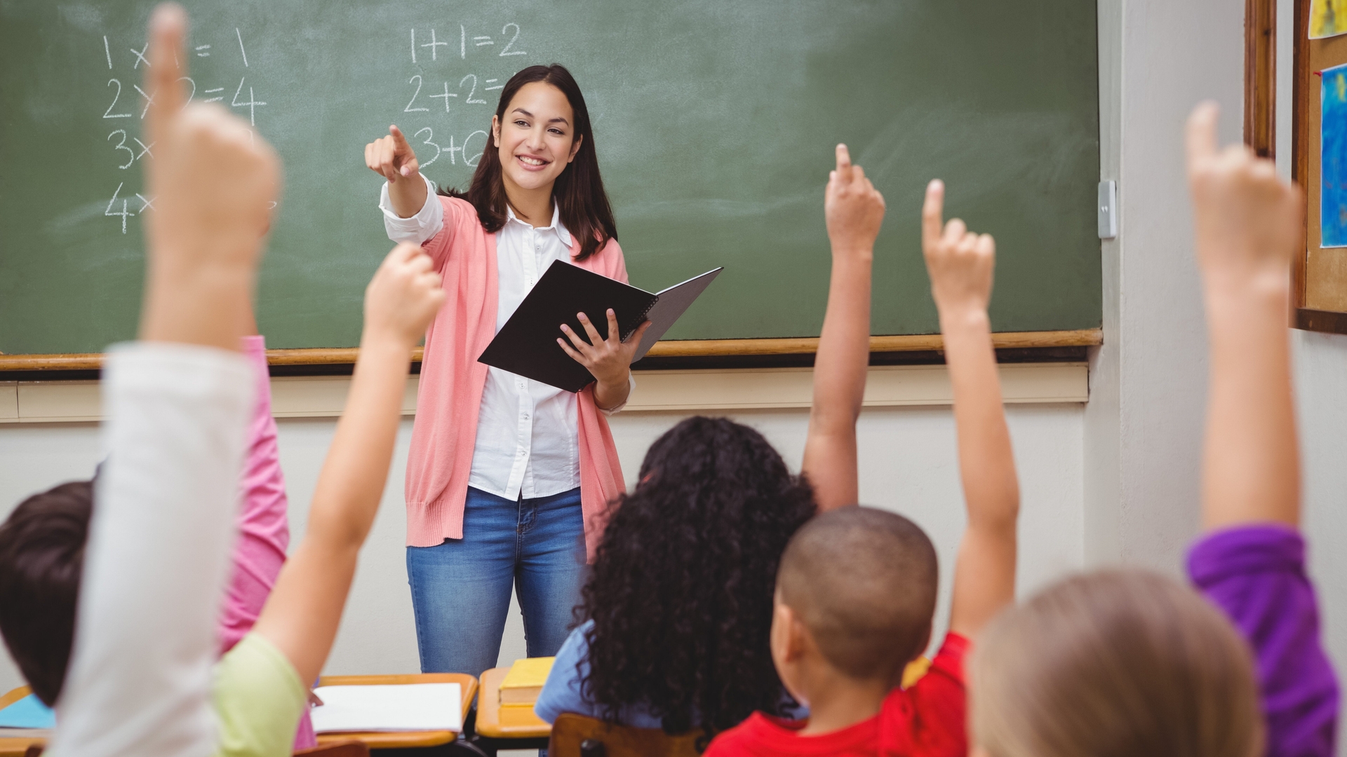 A teacher stands in front of a chalkboard and points at students with their hands raised.