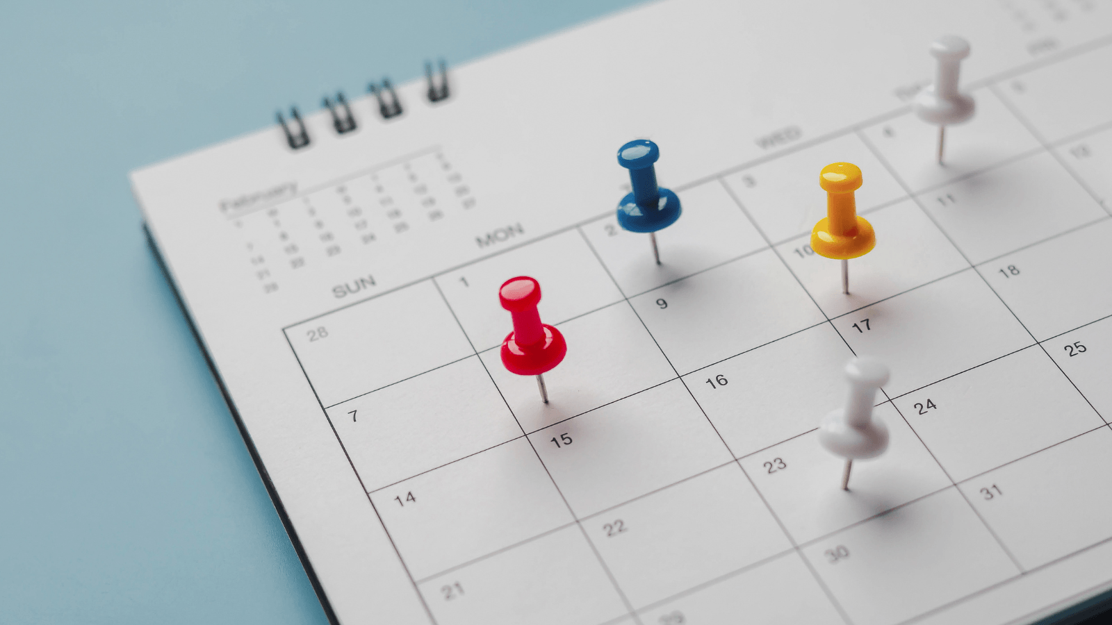 A calendar with pushpins marking specific dates in red, yellow, blue, and white on a spiral-bound page.