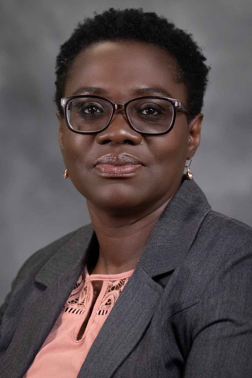 A woman wearing glasses, a gray blazer, and a peach blouse poses in front of a gray background.