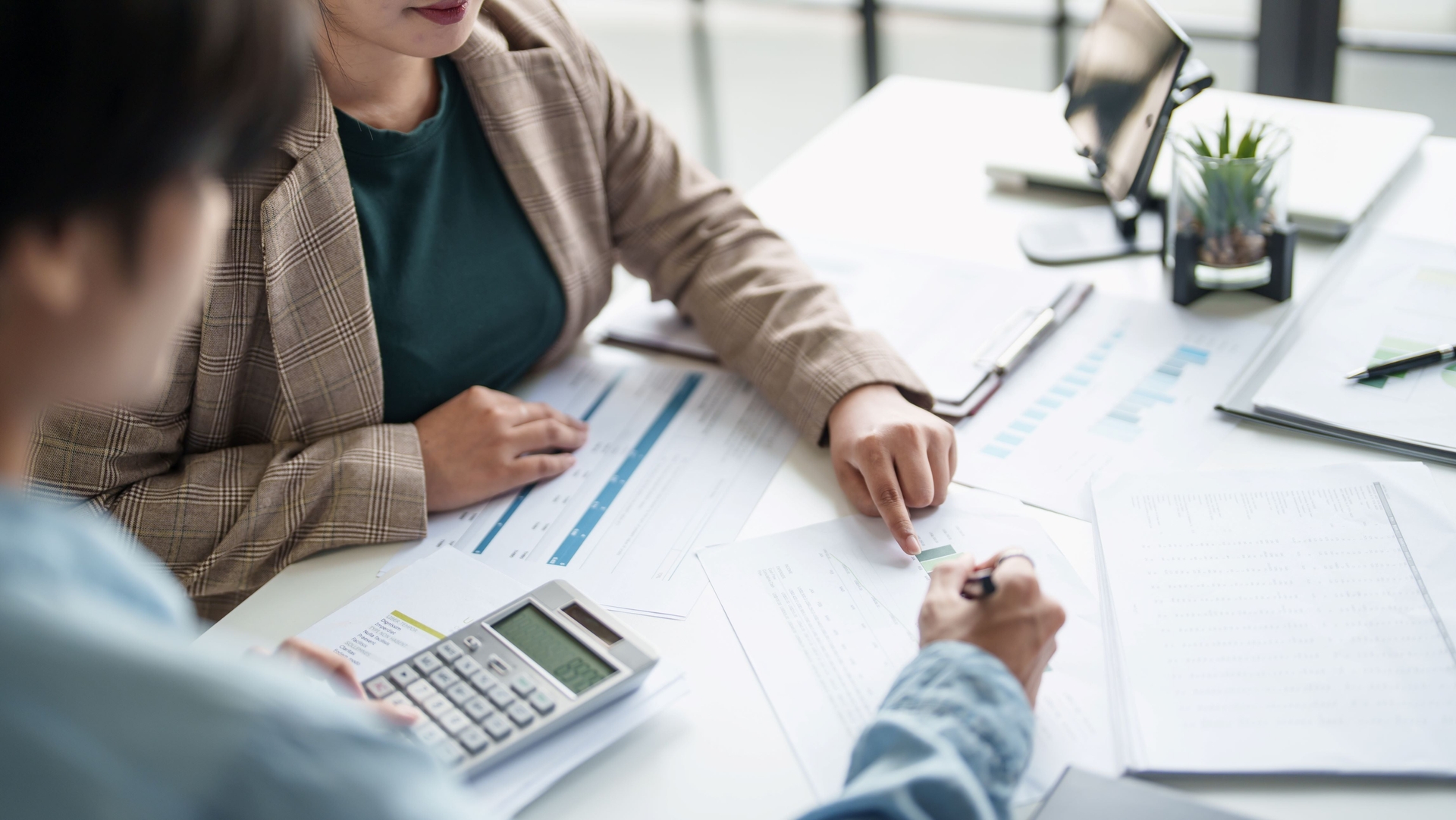 Two people review financial documents at a desk with charts, a clipboard, and a calculator, discussing figures and pointing to data on paper.