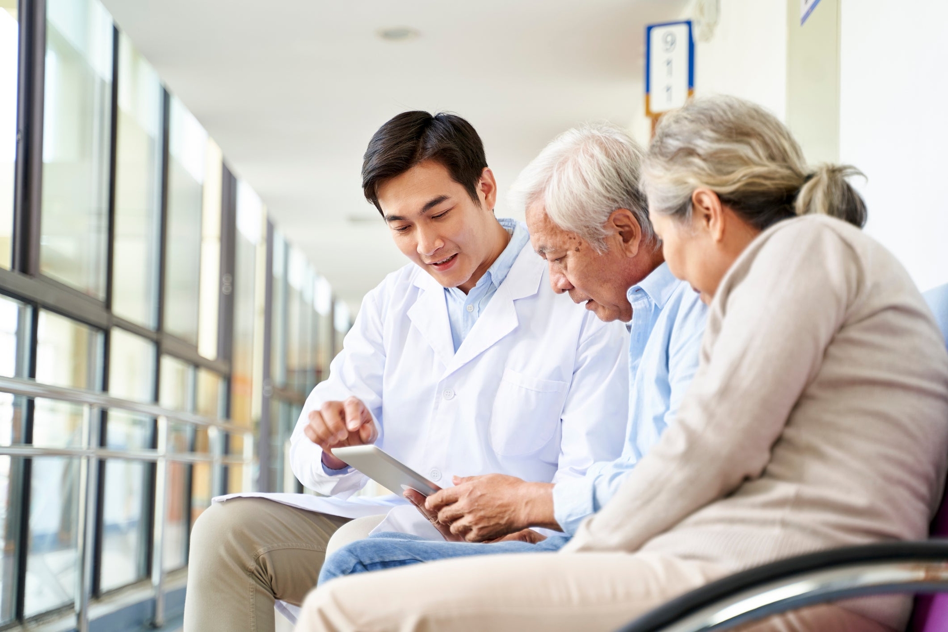A doctor shows information on a tablet to two older patients seated in a hallway, likely in a medical facility.