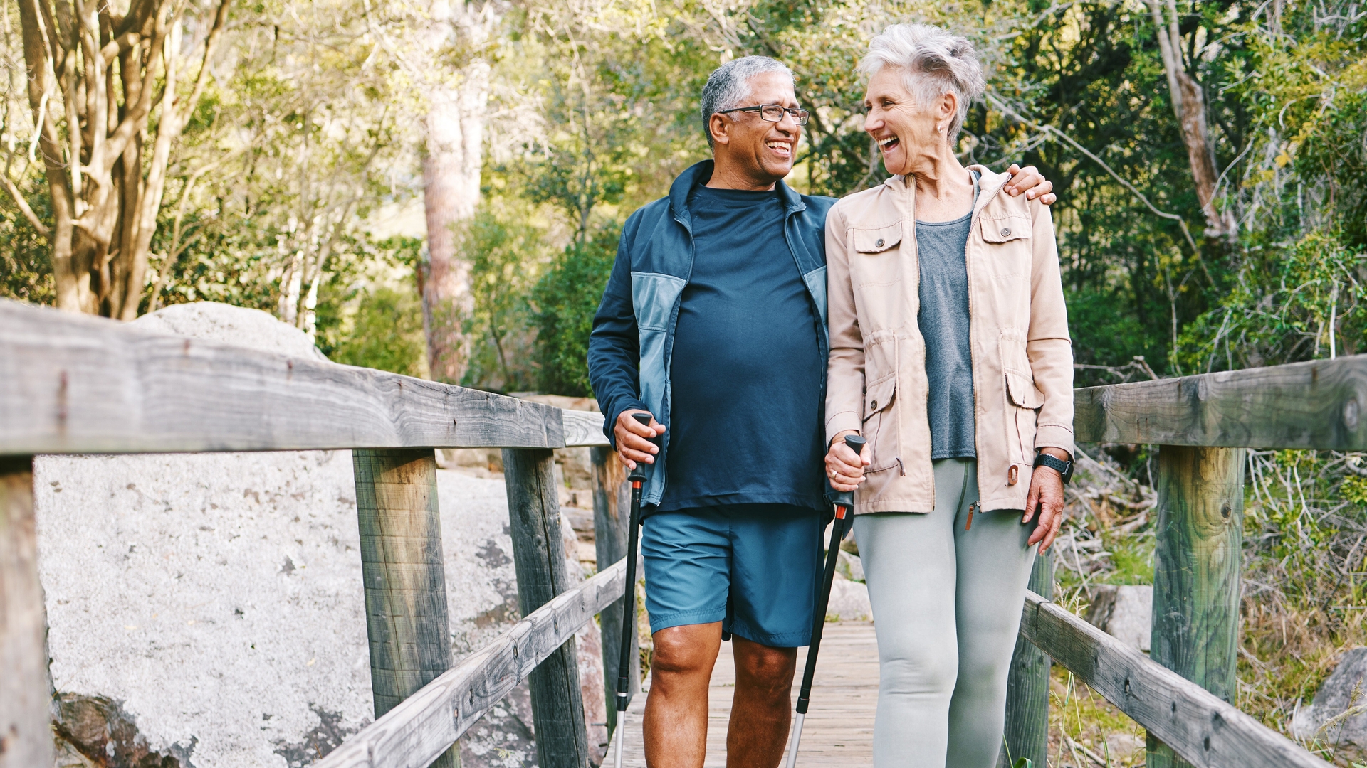 An older man and woman walk together on a wooden bridge in a forested area, smiling and holding hands.