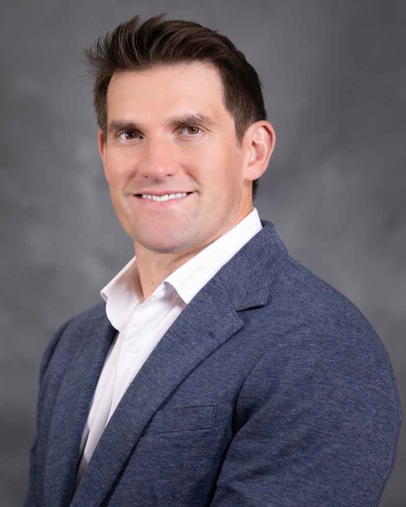 A man with short brown hair wearing a blue blazer and white shirt poses in front of a gray background.