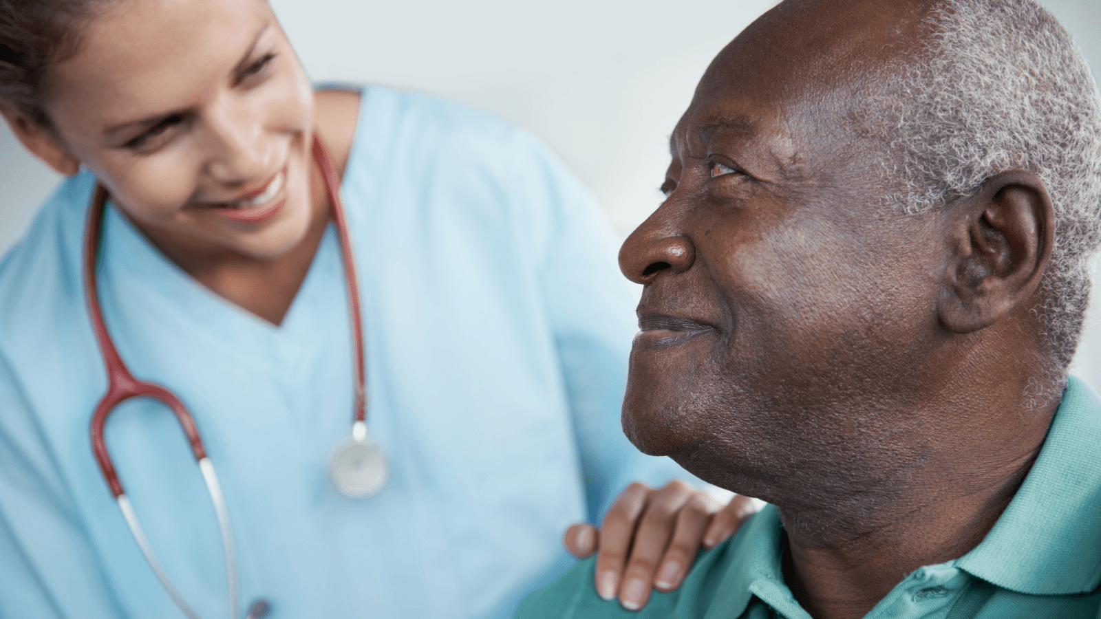 A healthcare worker with a stethoscope smiles and places a reassuring hand on the shoulder of an elderly man, who is smiling back.