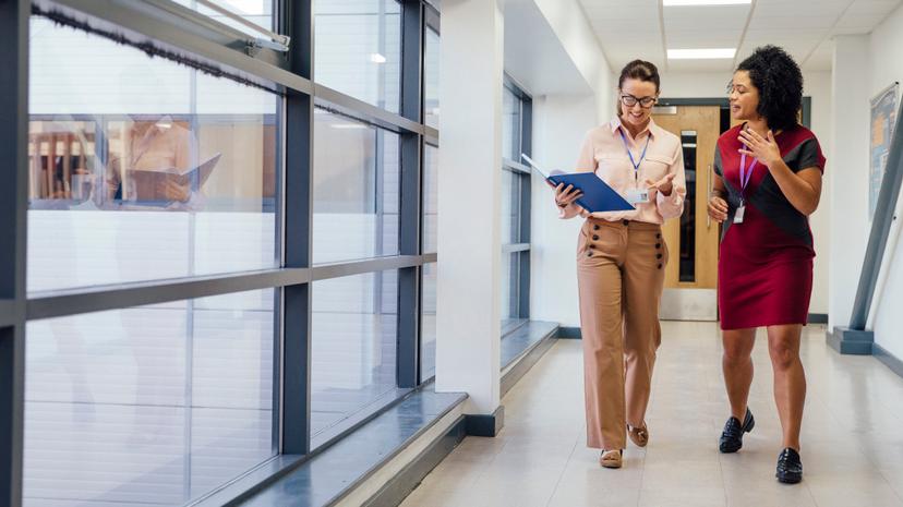 Two women walk down a hallway, one holding an open folder while they talk. Both are wearing ID lanyards and business attire. Large windows line one side of the corridor.