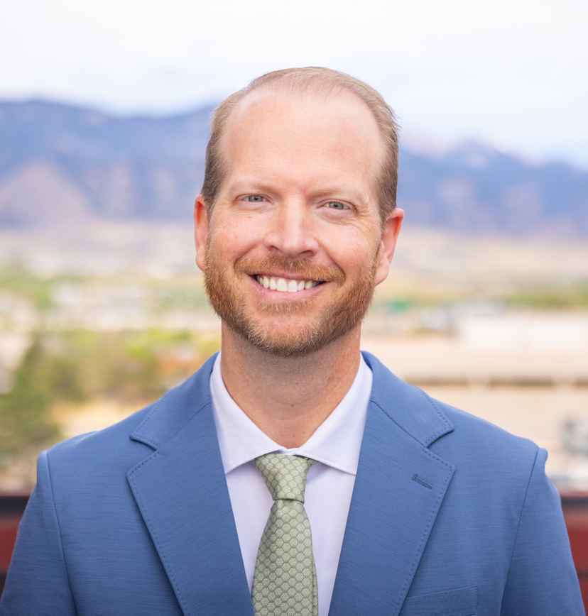 Man in a blue suit and green tie smiling outdoors with a blurred background of mountains and buildings.